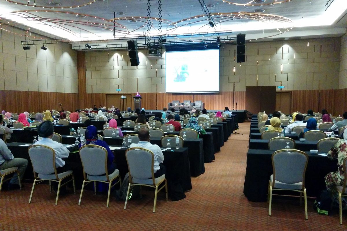 Delegates seated at rows of long tables in the ballroom, facing the stage and a large screen