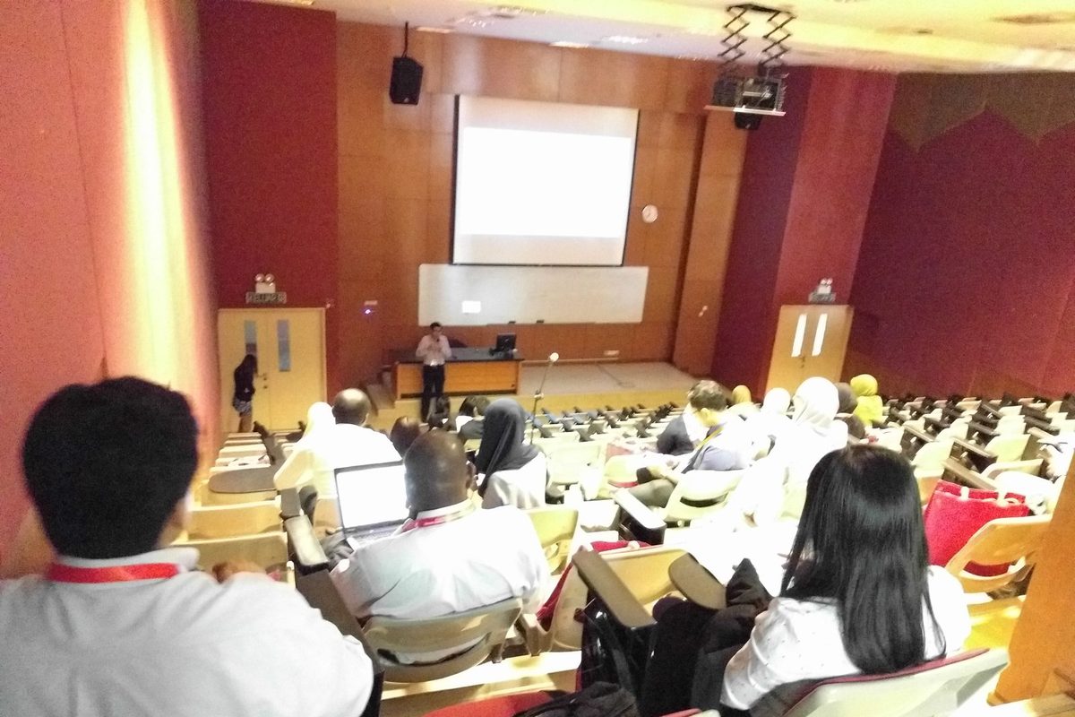 A presenter at the front of a lecture hall, with delegates seated in rows and a projection screen