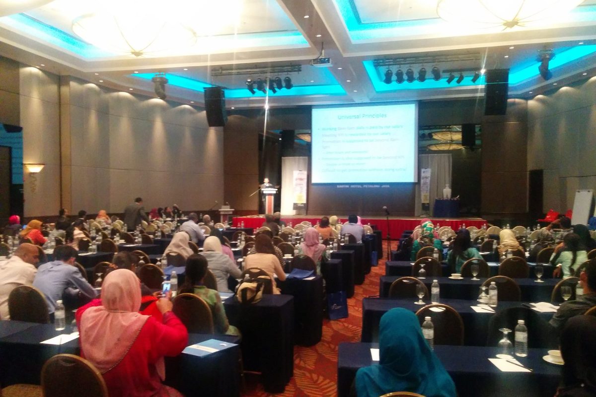 Delegates seated at long tables in a hotel ballroom, facing the stage and a projected lecture slide