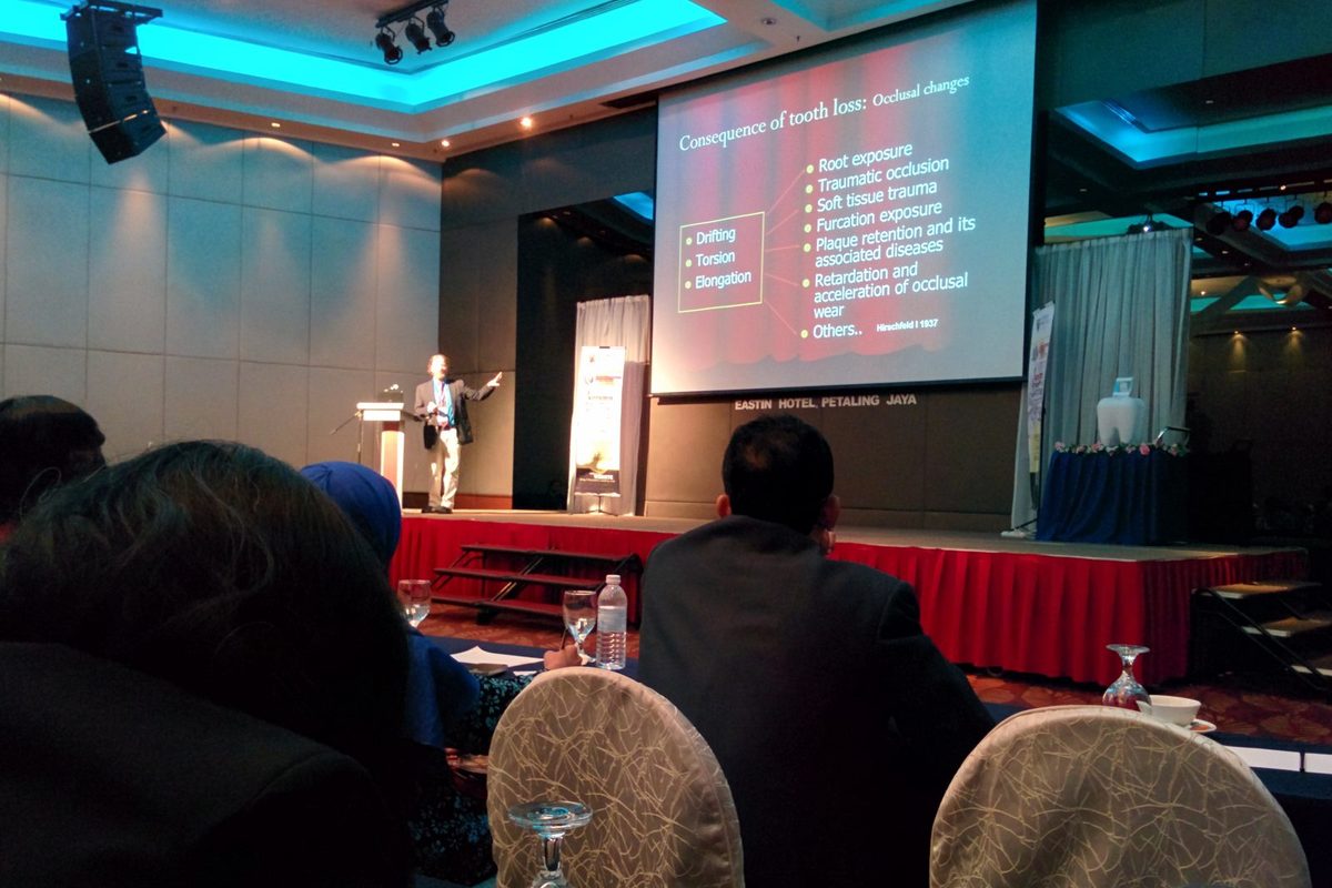 A speaker on stage presents a slide on the consequences of tooth loss to a seated audience
