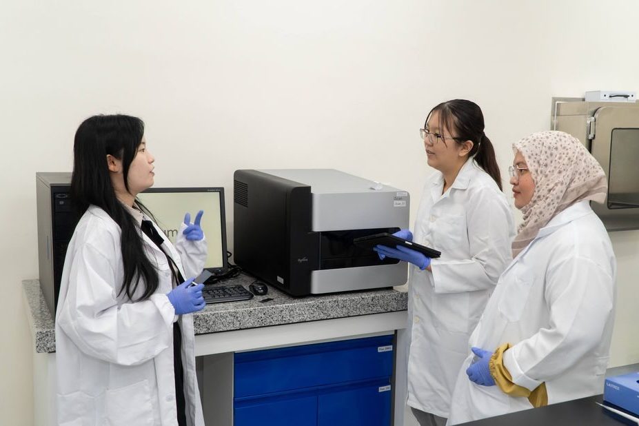 Three scientists in lab coats talking beside a laboratory instrument, one of them holding a tablet