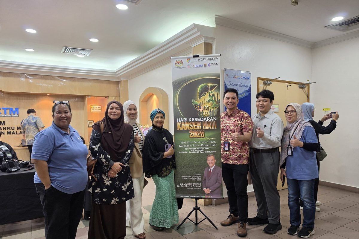 Seven people standing beside the Hari Kesedaran Kanser Hati 2026 banner in the auditorium foyer of Hospital Canselor Tuanku Muhriz