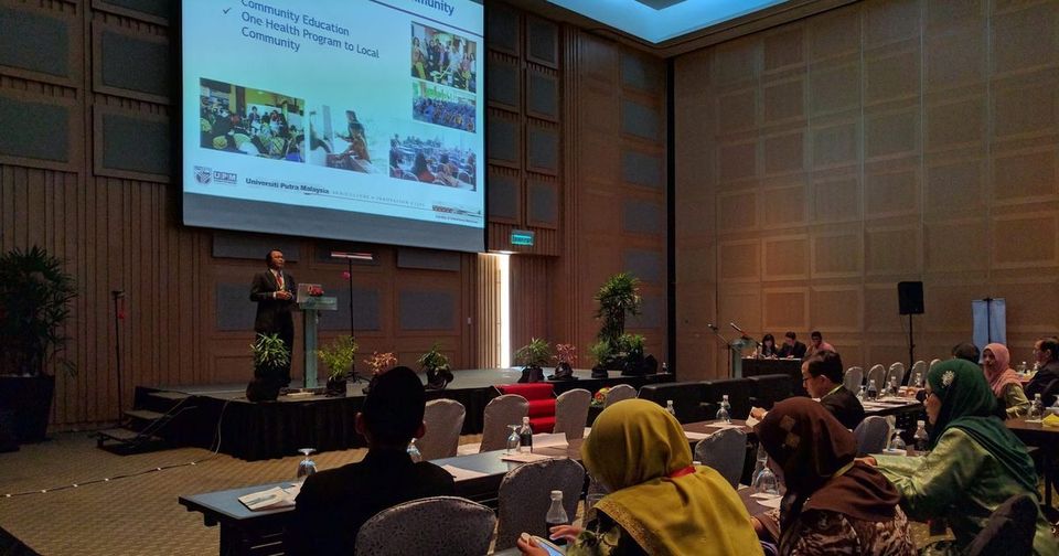 A speaker presents to delegates seated at long tables in a conference hall, with slides on a large screen