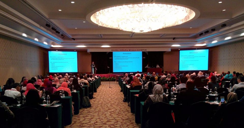 Full conference hall with delegates at long tables facing a speaker and three screens showing a slide on antioxidants