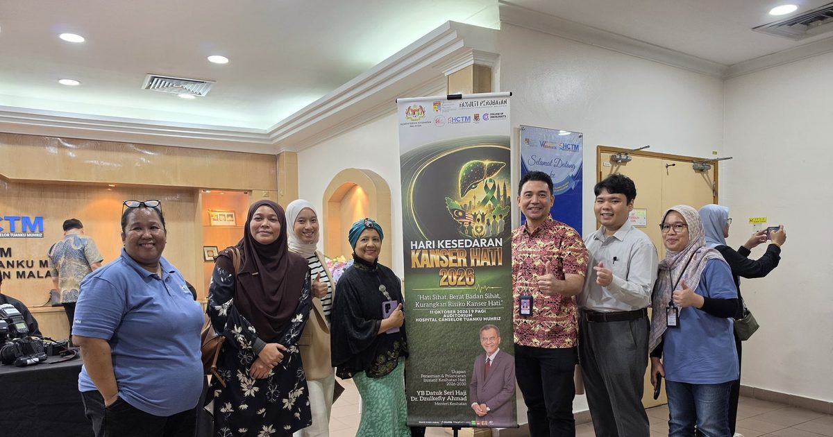 Seven people standing beside the Hari Kesedaran Kanser Hati 2026 banner in the auditorium foyer of Hospital Canselor Tuanku Muhriz