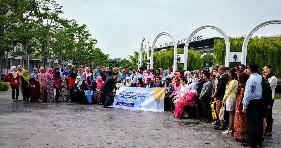 Conference participants in a large group photo outdoors in front of white arches, holding the conference banner