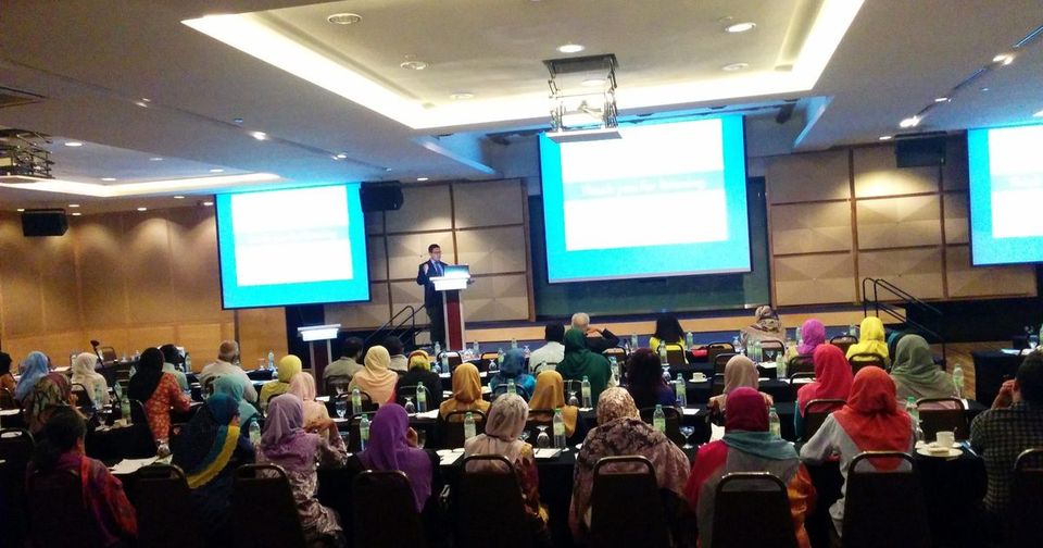 Delegates seated at long tables in a hotel ballroom, facing a speaker at a lectern and three large screens
