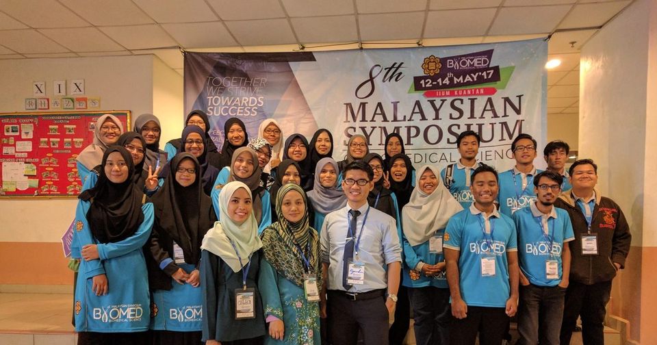 A large group, many in blue symposium T-shirts, poses in front of the 8th Malaysian Symposium of Biomedical Sciences banner