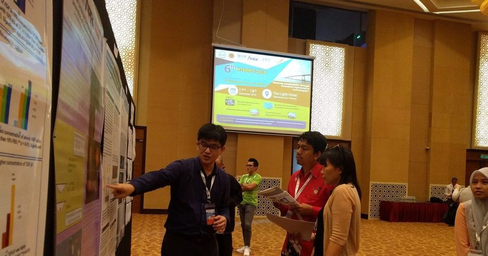 A man points to a research poster as two visitors listen, with the meeting slide on a screen behind