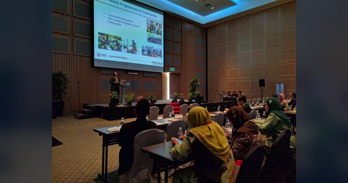 A speaker presents to delegates seated at long tables in a conference hall, with slides on a large screen