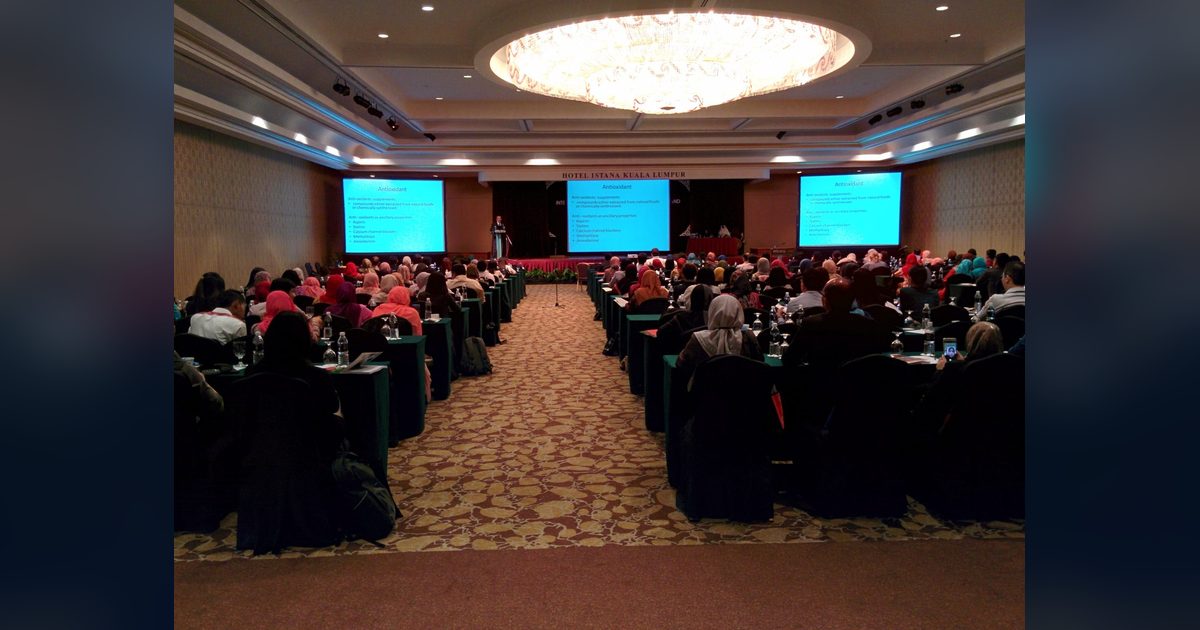 Full conference hall with delegates at long tables facing a speaker and three screens showing a slide on antioxidants