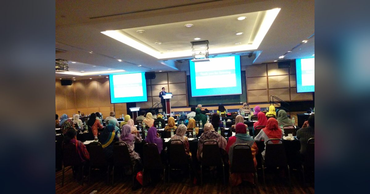 Delegates seated at long tables in a hotel ballroom, facing a speaker at a lectern and three large screens