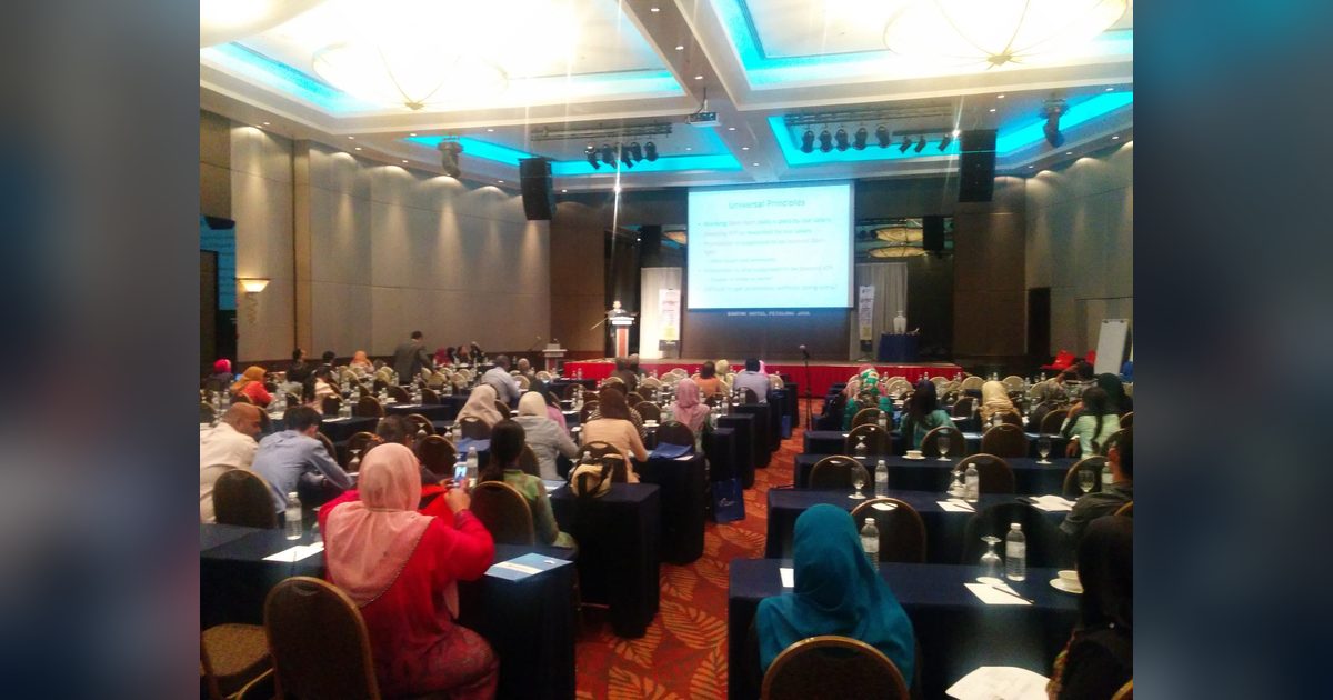 Delegates seated at long tables in a hotel ballroom, facing the stage and a projected lecture slide